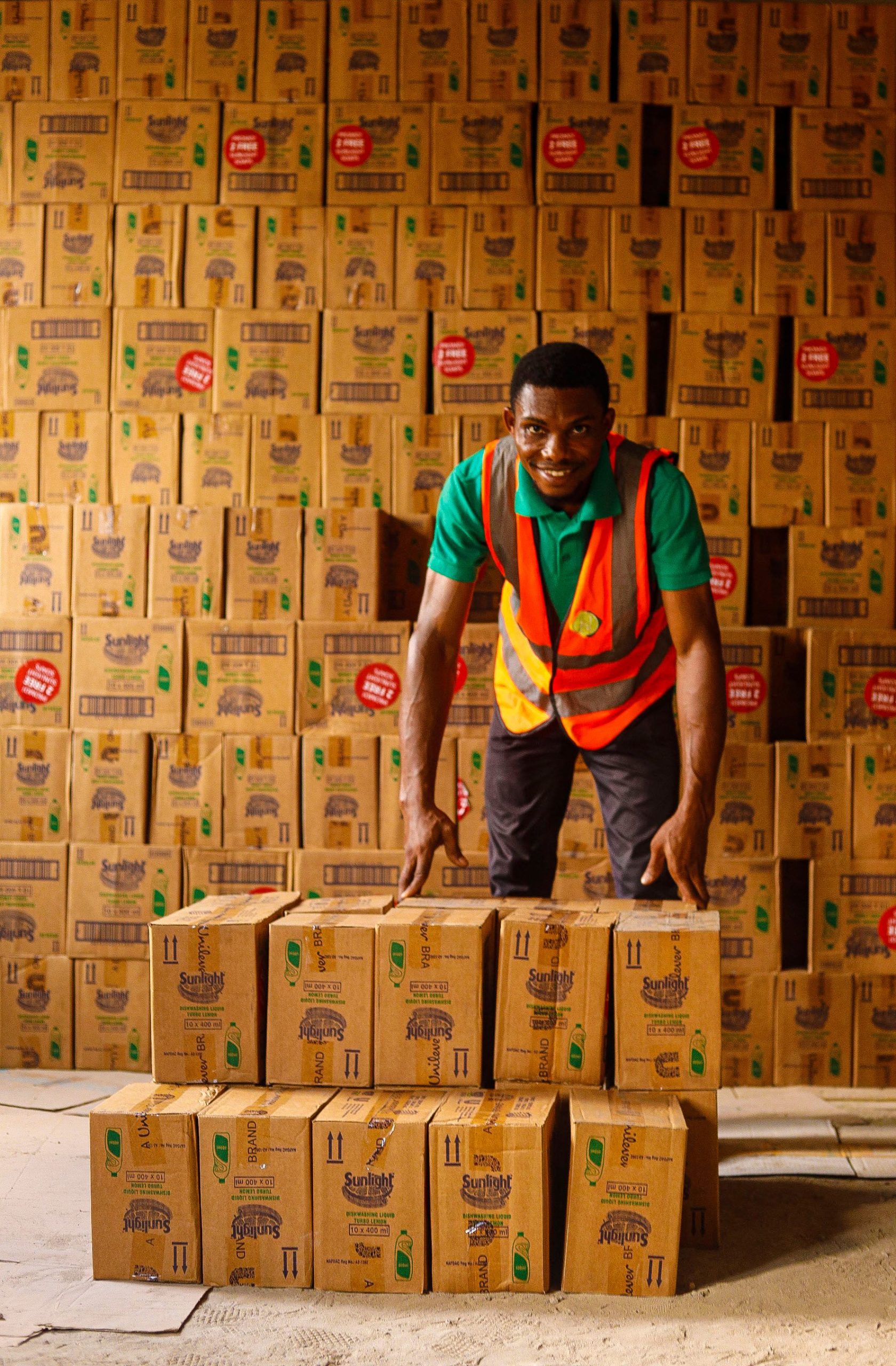 About Smiling warehouse worker organizing stacks of boxes with a reflective vest indoors.